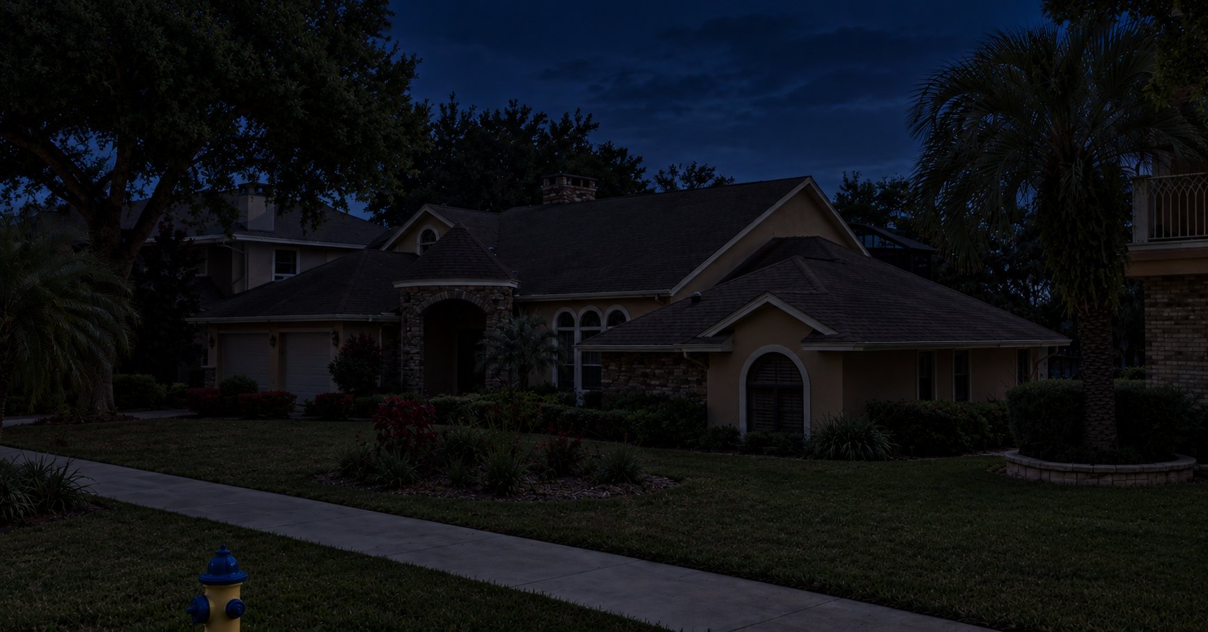 A stucco home at night with no exterior lighting: the roofline, entry arch, palms, and garden beds are all in darkness
